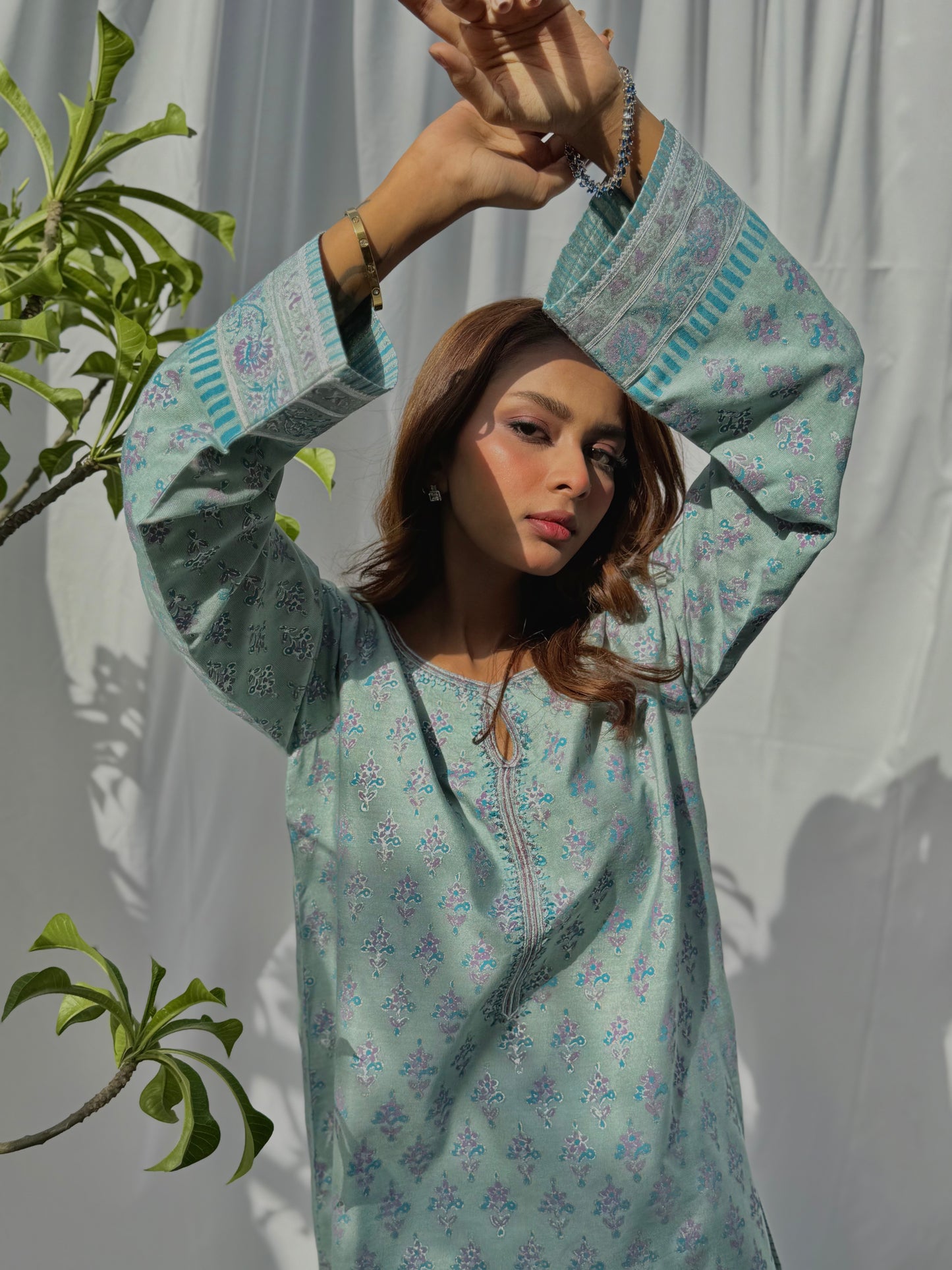 A woman wearing a light blue floral dress with a plant to the left on a white background