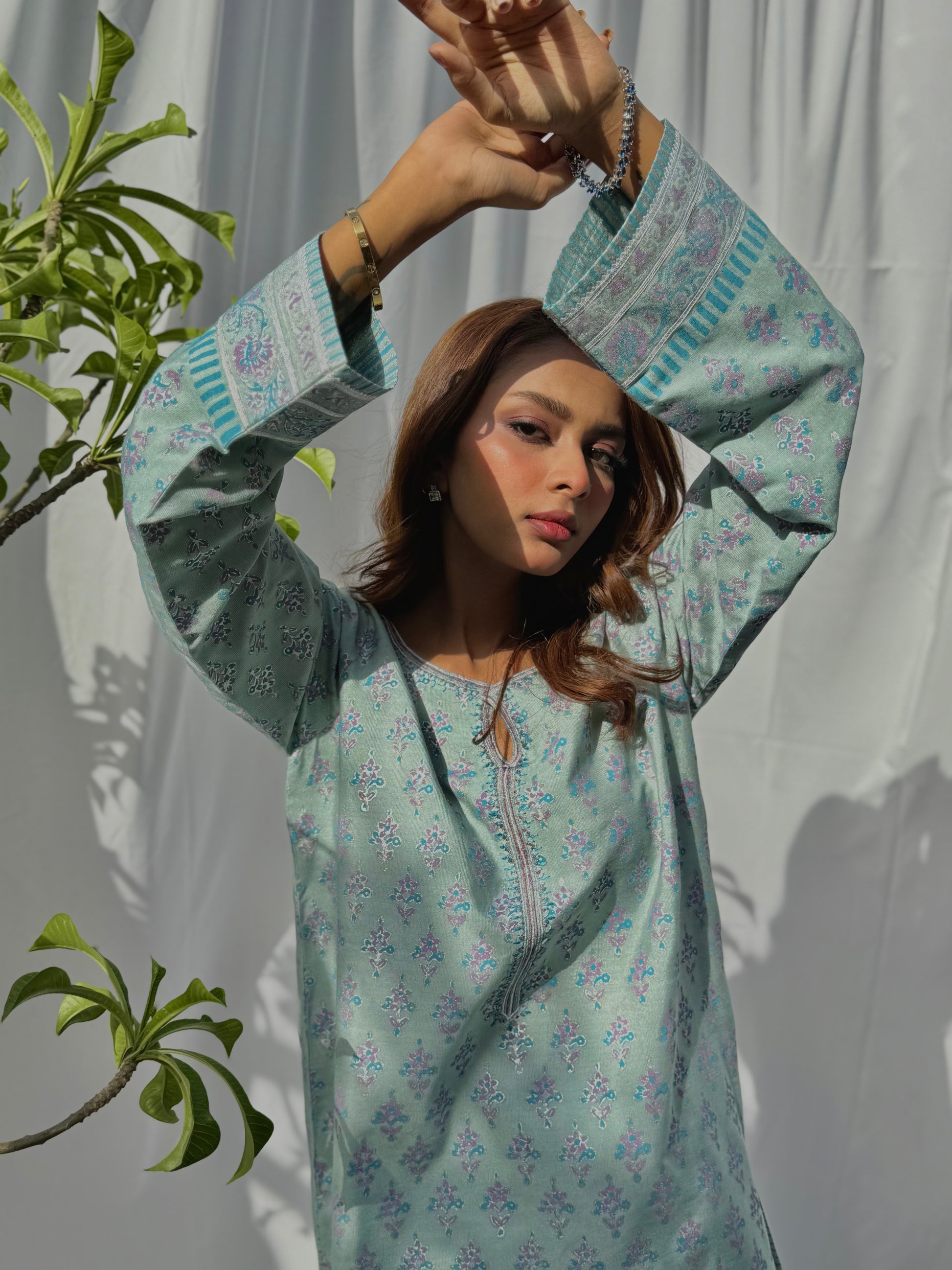 A woman wearing a light blue floral dress with a plant to the left on a white background